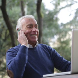 Elderly man joyfully using a laptop and phone outdoors, embracing technology.